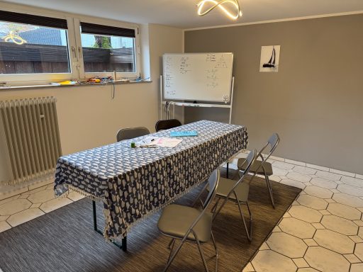 Ein Raum mit einem Tisch und mehreren Stühlen, einer Tafel und einem Teppich.